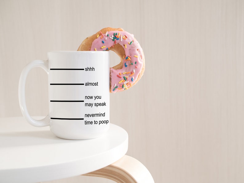 K&ouml;nnte beinhalten: Eine wei&szlig;e Keramiktasse mit schwarzen Linien und dem Text "shhh, fast, jetzt kannst du sprechen, vergiss es Zeit zum Kacken". Ein rosafarbener, glasierter Donut mit bunten Streuseln befindet sich teilweise in der Tasse.