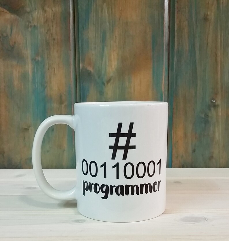 K&ouml;nnte beinhalten: Wei&szlig;er Keramikbecher mit schwarzem Henkel. Der Becher hat ein schwarzes Hashtag-Symbol, Bin&auml;rcode und das Wort "Programmierer" in Schwarz aufgedruckt. Der Hintergrund ist eine Holzoberfl&auml;che.