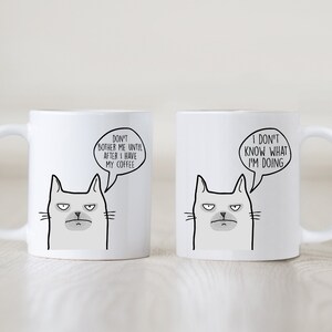 May include: Two white ceramic mugs with black cat illustrations and humorous text. The mug on the left says "Don't bother me until after I have my coffee." The mug on the right says "I don't know what I'm doing."