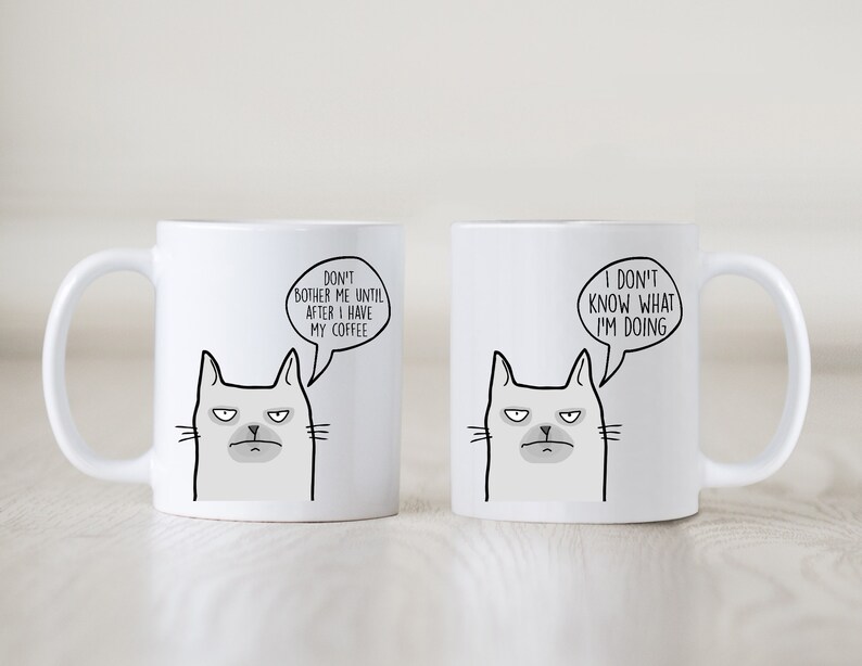 May include: Two white ceramic mugs with black cat illustrations and humorous text. The mug on the left says "Don't bother me until after I have my coffee." The mug on the right says "I don't know what I'm doing."