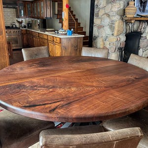 May include: A large, round, wooden dining table with a natural wood finish. The table is surrounded by chairs with brown leather upholstery.