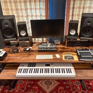 May include: A home recording studio setup featuring a large wooden desk with a keyboard, computer monitor, speakers, and audio mixing equipment. The desk sits on a patterned rug, with curtains in the background.