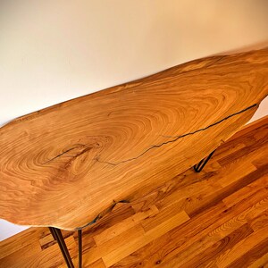 May include: A natural wood console table with a live edge top and black hairpin legs. The table has a light brown wood grain and a natural finish.