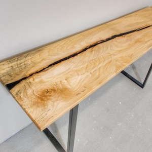 May include: A rectangular wooden console table with a light brown finish and a dark black line running down the center. The table has a black metal frame. The table is approximately 60 inches long.