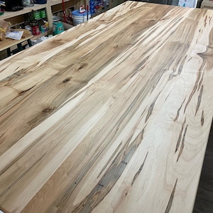 May include: A close-up of a light brown wood table top with a natural wood grain pattern. The wood has a light brown color with darker brown streaks throughout. The table top is smooth and polished.
