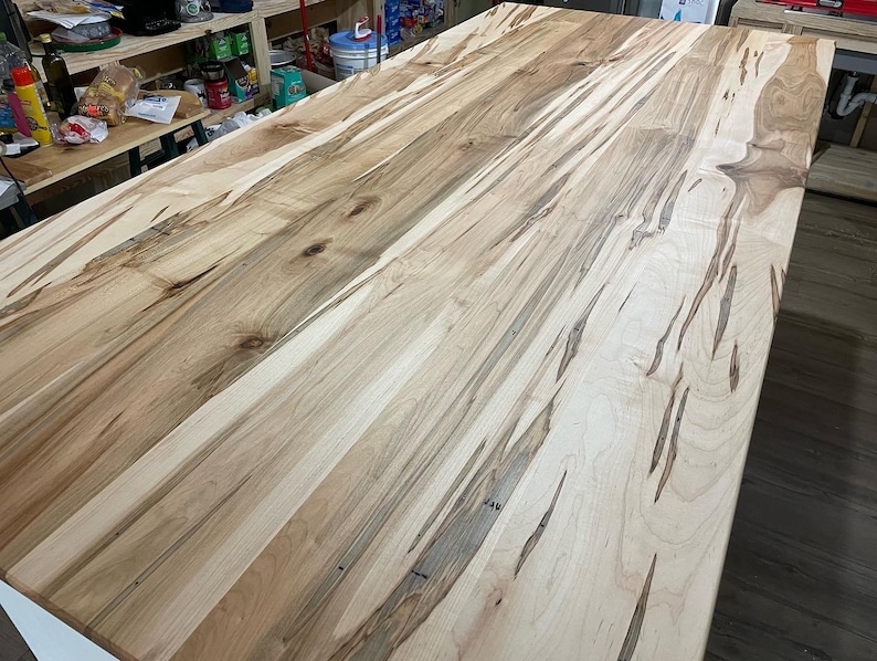 May include: A close-up of a light brown wood table top with a natural wood grain pattern. The wood has a light brown color with darker brown streaks throughout. The table top is smooth and polished.