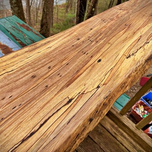 May include: A close-up of a long, wooden plank with a natural, live edge. The wood has a rich, brown color with streaks of lighter wood running through it. The plank is smooth and polished, and it appears to be a high-quality piece of wood.
