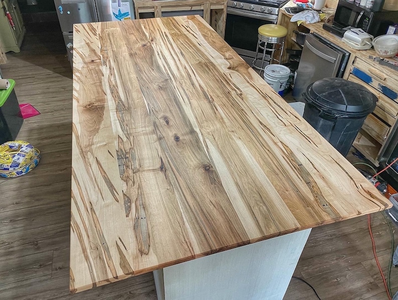 May include: A close-up of a large, light brown wooden kitchen island countertop with a natural wood grain pattern. The countertop is made of maple wood and has a smooth, polished finish.