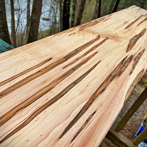 May include: A close-up of a long, wooden board with a natural wood finish. The board has a unique grain pattern with dark brown streaks and knots. The board is likely made of maple or cherry wood.