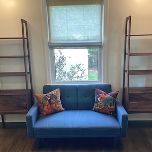 May include: Two wooden ladder shelves with a drawer at the bottom, flanking a blue sofa with two patterned pillows. The sofa is in front of a window with a white roman shade.