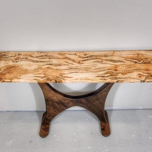 May include: A handcrafted wooden console table with a light-colored, patterned top and a dark brown, curved base. The table's top is approximately 60 inches long, showcasing natural wood grain. The base has a unique, artistic design.