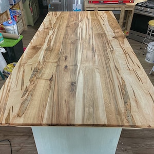 May include: A close-up view of a light wood kitchen island countertop with a natural wood grain pattern. The countertop is made of multiple pieces of wood that have been joined together.