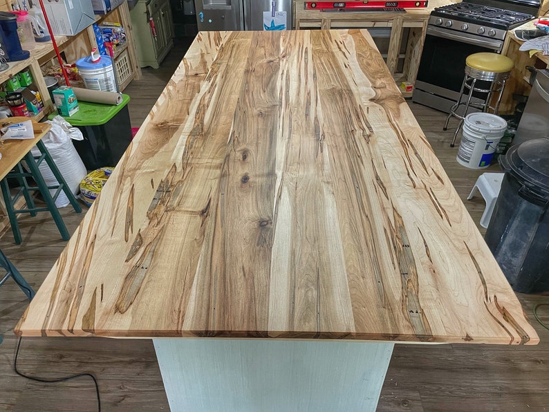 May include: A close-up view of a light wood kitchen island countertop with a natural wood grain pattern. The countertop is made of multiple pieces of wood that have been joined together.
