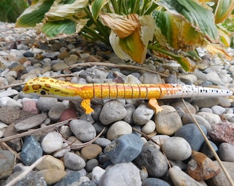 May include: A hand-painted wooden gecko lizard toy with yellow, orange, and white paint. The gecko is posed on a bed of small grey and brown stones.