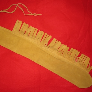 Puede incluir: Una bolsa de cuero larga y de color marrón claro con un borde de flecos y una correa de cuero. La bolsa está sobre una superficie roja.