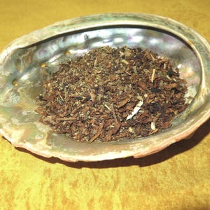 Op de afbeelding: Een close-up van een bruin en groen kruidenmengsel in een iriserende abalone-schelp. De schelp ligt op een gele ondergrond.