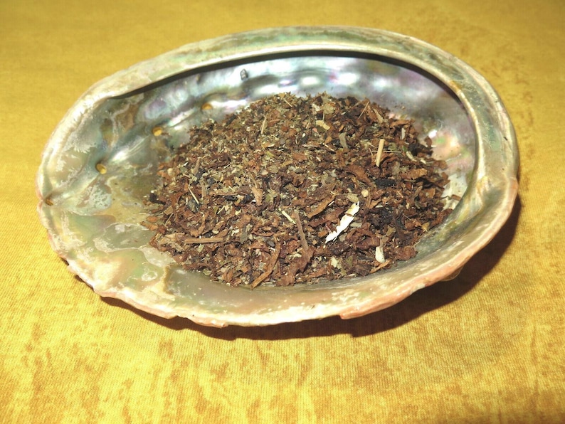 Op de afbeelding: Een close-up van een bruin en groen kruidenmengsel in een iriserende abalone-schelp. De schelp ligt op een gele ondergrond.