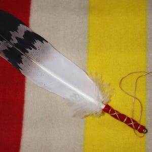 May include: A large feather with a black and white pattern, a red and gold wrapped handle, and a beaded cord. The feather is set against a backdrop of red, yellow, and cream-coloured fabric.