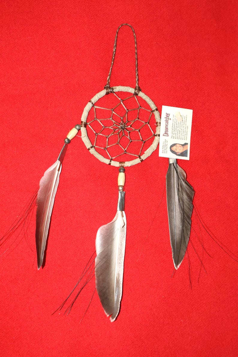 May include: A brown and white dreamcatcher with three feathers hanging from it. The dreamcatcher is on a red background. The card attached to the dreamcatcher says "Dreamcatcher".