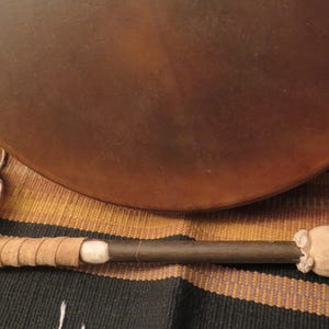 May include: A brown, circular drum with a matching mallet. The mallet features a dark wooden handle, a tan felt head, and tan leather wrapping. The drum is partially visible, with a smooth, dark brown surface. The background is a patterned textile.