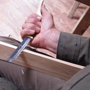 Peut inclure: Une personne utilise un couteau à sculpter le bois pour façonner un morceau de bois. Le bois est brun clair et le couteau est argenté avec un manche brun.