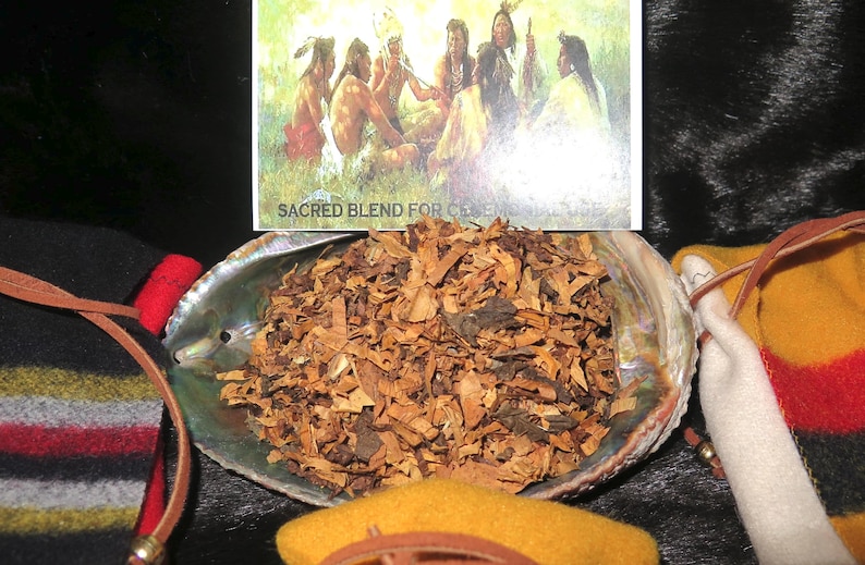 Op de afbeelding: Een close-up van een schelp gevuld met gedroogde kruiden en houtsnippers. De schelp ligt op een bruin-zwarte gestreepte deken. Een kaart met de tekst "Heilige mix voor ceremonie" is zichtbaar op de achtergrond.