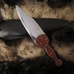 May include: A decorative knife with a polished steel blade and a dark wood handle. The handle has a circular design with inlaid white dots. The knife rests on a black sheath, against a textured brown and black background.