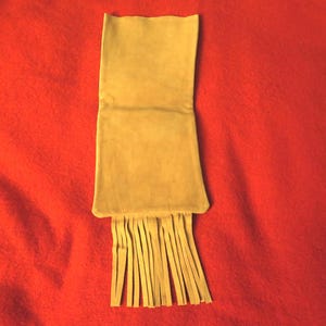 May include: A light brown suede pouch with a fringe trim. The pouch is folded in half and has a rectangular shape.