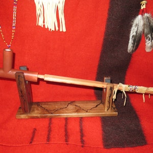 Puede incluir: Un soporte de madera con una pipa de arcilla roja y una boquilla de madera. El soporte está decorado con un patrón de veta de madera oscura. La pipa está decorada con cuentas y plumas.