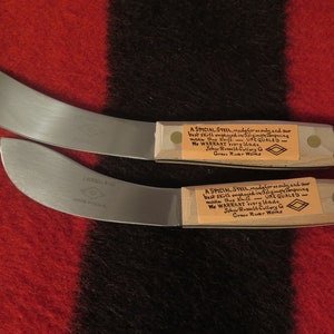 Könnte beinhalten: Zwei Messer mit Holzgriffen und Klingen aus rostfreiem Stahl. Die Klingen sind gebogen und haben einen leichten Aufwärtsschwung. Die Griffe sind aus Holz und haben einen Messingniet. Die Messer haben ein Etikett am Griff, auf dem steht "A SPECIAL STEEL made for us only and our best skill employed in forging tempering make thus Knife - UNEQUALED - We WARRANT every Made John Russell Cutlery Co Green River Works".