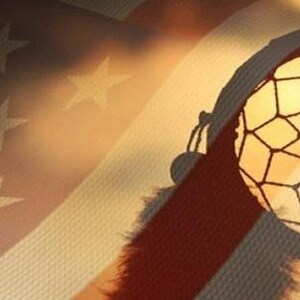 May include: A dreamcatcher with a brown leather cord and white feathers hangs against a blurred background of an American flag.