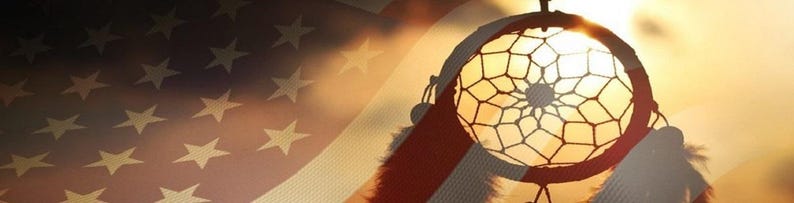 May include: A dreamcatcher with a brown leather cord and white feathers hangs against a blurred background of an American flag.