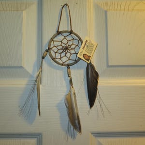 May include: A brown and white dreamcatcher with feathers hanging from it. The dreamcatcher is hanging on a white door. The tag attached to the dreamcatcher reads "Dreamcatcher".