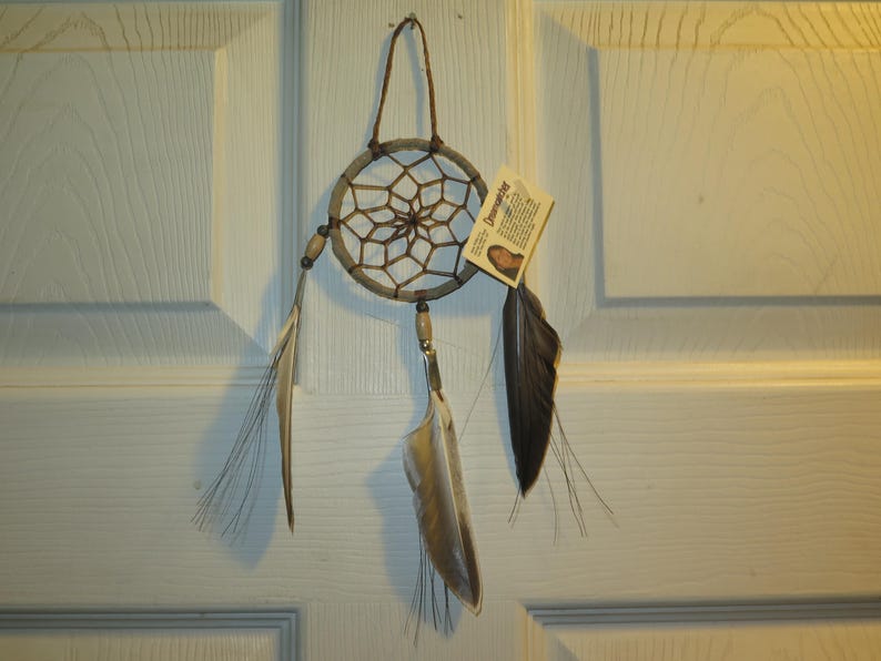 May include: A brown and white dreamcatcher with feathers hanging from it. The dreamcatcher is hanging on a white door. The tag attached to the dreamcatcher reads "Dreamcatcher".
