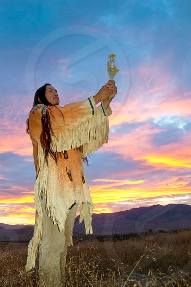 Op de afbeelding: Een indiaanse man in traditionele kleding houdt een bosje wilde bloemen vast voor een kleurrijke zonsondergang. Hij staat in een veld met hoog gras.