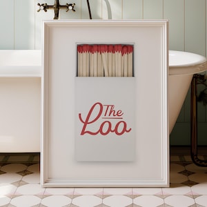 May include: A framed vertical print featuring a white matchbox with red-tipped matches. The box displays the red text "The Loo" in a cursive script font. The print is presented in a simple white frame, showcasing a clean and graphic design.