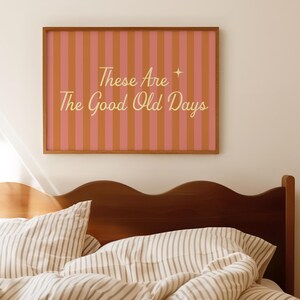 May include: A framed print with a pink and orange striped background and the text "These Are The Good Old Days" in gold lettering. The print is hanging above a bed with striped bedding and a wooden headboard.