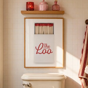 May include: A framed wall art print featuring a white matchbox with red-tipped matches and the red text The Loo. The print is displayed above a white toilet in a tiled bathroom, with a wooden shelf holding a red candle, a pink jar, and a reed diffuser.