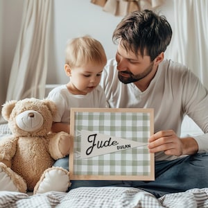 May include: A light wooden framed sign with a green and white checkered background. The sign has a white pennant with the name "Jude" in black script and the name "Dylan" in a smaller font. A teddy bear sits next to the sign.