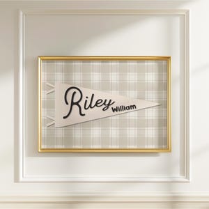 May include: A framed pennant with the name "Riley William" in black script. The pennant is cream-colored and set against a plaid background. The frame is gold, and the wall is white.