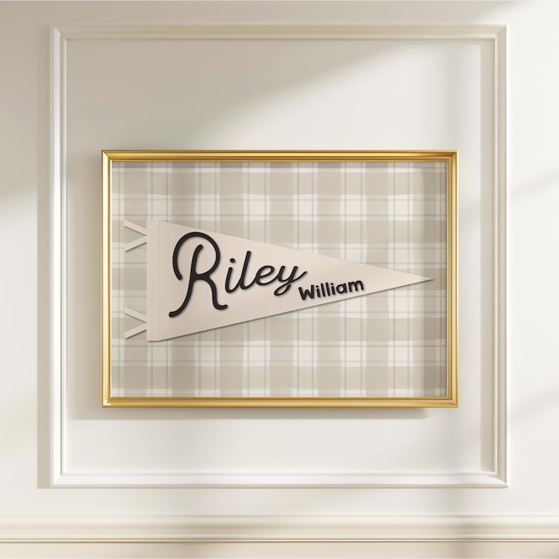 May include: A framed pennant with the name "Riley William" in black script. The pennant is cream-colored and set against a plaid background. The frame is gold, and the wall is white.
