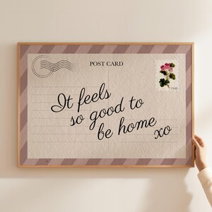 May include: A framed postcard print with the text "It feels so good to be home xo" in elegant script. The design includes a postage stamp with a floral illustration and a postmark graphic. The frame has a light wood finish.