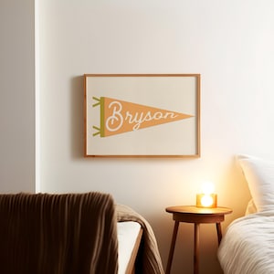 May include: A framed print of a peach-colored pennant with the word "Bryson" written in white cursive. The pennant is hanging on a white wall above a wooden nightstand with a lamp.