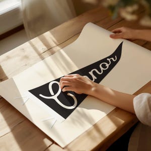 May include: A black and white pennant banner with the name "Connor" written in a cursive font. The banner is being placed on a white surface.