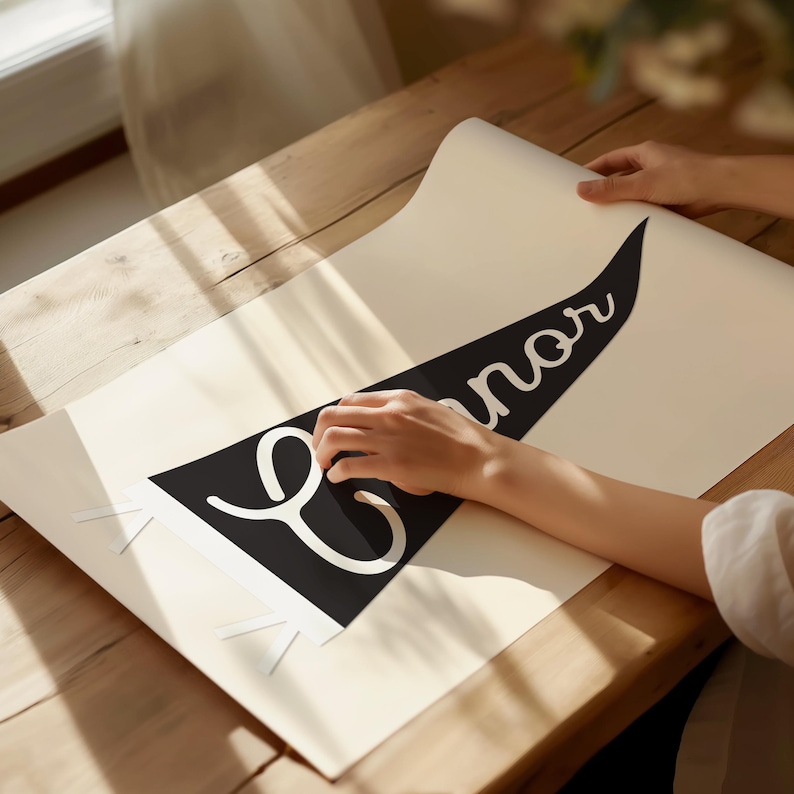 May include: A black and white pennant banner with the name "Connor" written in a cursive font. The banner is being placed on a white surface.