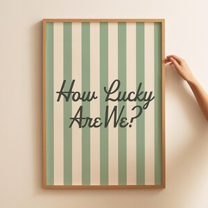 May include: A framed print with a light brown wooden frame. The print features vertical stripes in alternating shades of green and cream. The text "How Lucky Are We?" is written in a dark green, cursive font.
