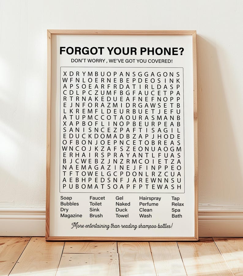 May include: A black and white printable word search puzzle with the words "Forgot your phone? Don't worry, we've got you covered!" at the top. The puzzle includes words related to bathroom items, such as soap, bubbles, faucet, toilet, gel, naked, hairspray, perfume, tap, relax, dry, sink, duck, clean, spa, magazine, brush, towel, and wash. The puzzle is framed in a light wood frame.