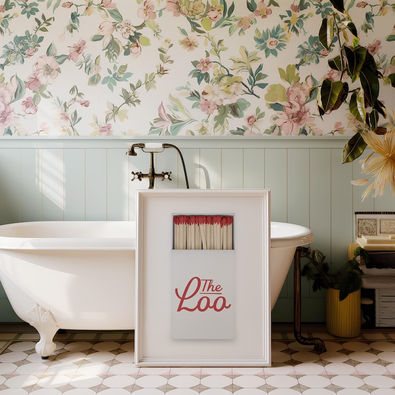 May include: A framed vertical print featuring a white matchbox with red-tipped matches. The box displays the red text "The Loo" in a cursive script font. The print is presented in a simple white frame, showcasing a clean and graphic design.