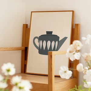 May include: A framed print of a minimalist illustration of a blue teapot with white leaf-shaped designs on a cream background.