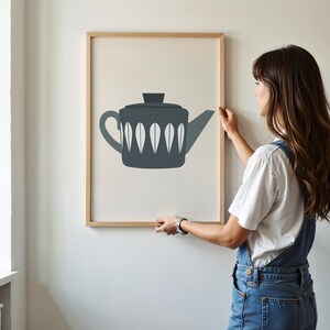 May include: A framed print of a minimalist illustration of a gray teapot with white leaf-shaped details against a cream background.